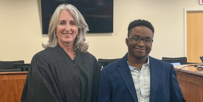 A judge in a black robe standing beside a person in a suit inside a courtroom.