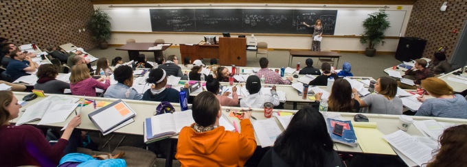 a view of a classroom with students taken from behind looking towards the chalkboard and professor. 
