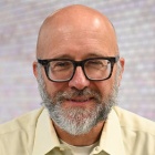 Man wearing glasses, shirt, blue pants, standing in front of a large glass window next to a table and blue chair.