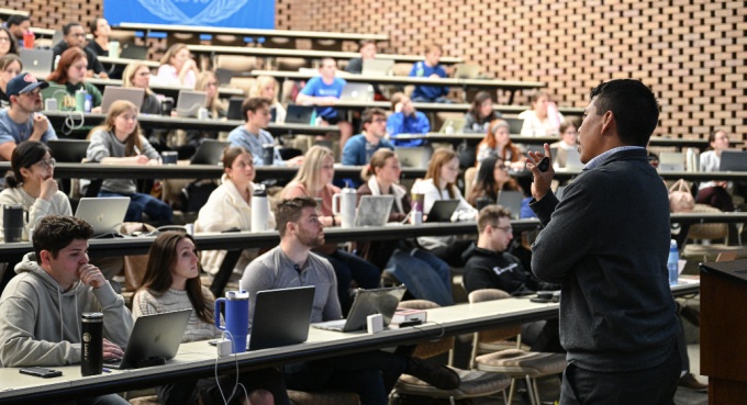 A professor standing in front of a classroom with several rows of students sitting at tables with books and laptops, listening to a lecture.