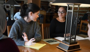 students sitting at a library table, talking to a professor.