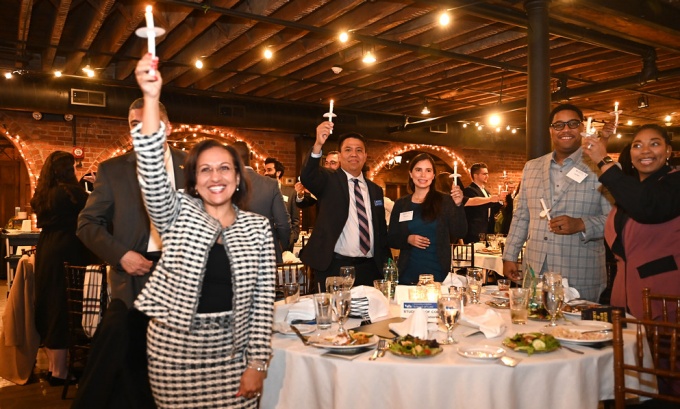 A group of peope attending an event, standing around tables, holding up lit candles. 