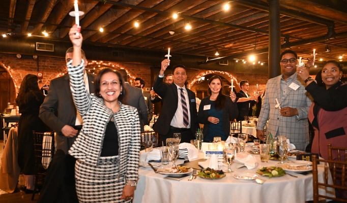 A group of peope attending an event, standing around tables, holding up lit candles. 