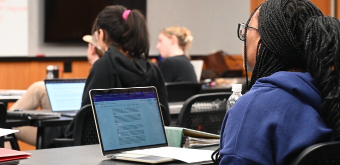 A classroom scene with several students seated at tables using laptops. The focus is on a student in the foreground wearing glasses and working on a laptop that displays a document. Other students sit at separate tables facing the front of the room, where a large screen is mounted on the wall.