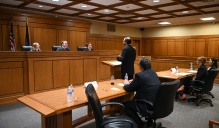 Courtroom scene with a panel of three judges seated behind a wooden bench while an attorney stands and speaks at a podium. Additional attorneys sit at tables facing the judges, with water bottles and papers arranged on the desks.