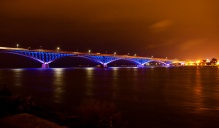 Nighttime view of a long bridge illuminated with vibrant blue lights, reflecting off the dark water below, with an orange-glowing sky and distant city lights in the background.