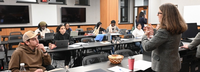 A classroom with multiple tables arranged in rows, where several people are seated with laptops, notebooks, and beverages in front of them. A person at the front of the room is speaking to the group, gesturing with their hands.