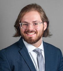 Young man wearing suit and tie, glasses and smiling.