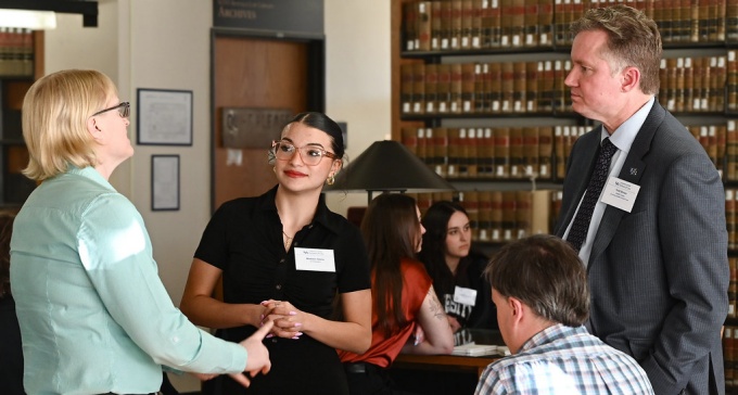 Group of three people standing in a library, two individuals listen as one speaks. 