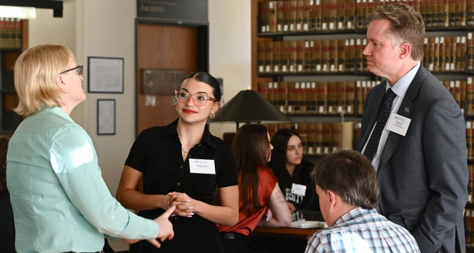 Group of three people standing in a library, two individuals listen as one speaks.