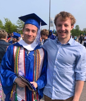 two men, one wearing graduation regalia, smiling outside.