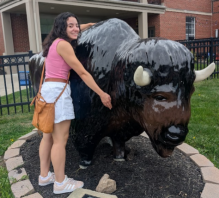 A person stands outdoors beside a large black and brown bison statue with white horns. They wear a pink sleeveless top, white shorts, white sneakers, and a tan crossbody bag. One arm is around the bison’s neck and the other rests on its side. The statue is set on a circular mulch bed bordered by bricks, with a red brick building and black metal fencing in the background.