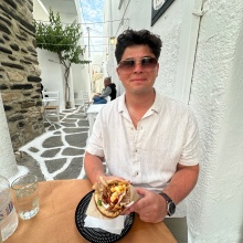 A person sits at an outdoor café table in a narrow, white‑washed alley with stone‑patterned pavement. They are holding a plate with a pita sandwich filled with vegetables and meat. Glasses of water and tableware are on the table, and other diners are seated further down the walkway.