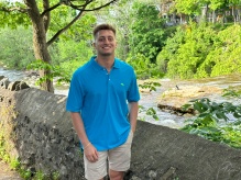 man smiling, standing outside next to a stream. 