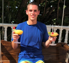 man sitting outside on a bench, holding two colorful drinks in each hand, smiling.