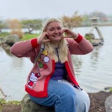 Young woman sitting next to a pond, wearing red Hello Kitty sweater, jeans and purple shirt.
