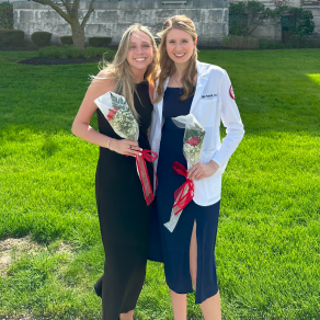 Two woman standing outside, next to each other, smiling and holding flowers. 