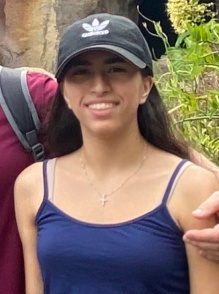 A person wearing a navy tank top and a black baseball cap stands outdoors in front of rocky terrain and greenery. Another person’s arm rests lightly on their shoulder from the side of the frame.