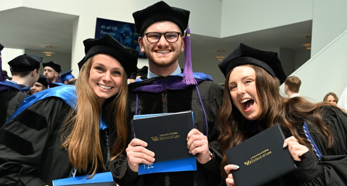 three recent graduates wearing their regalia and hold up their diplomas. 