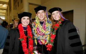 young adults standing, wearing flowery wreaths and graduation regalia. 