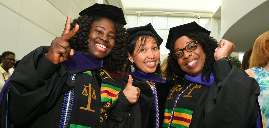 three women standing together wearing graduation regalia, smiling and giving a thumbs up gesture. 