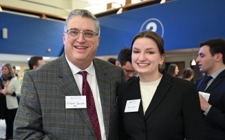 Professionally dressed man and woman standing together, smiling, attending an event in a large open space, with several people standing and talking. 