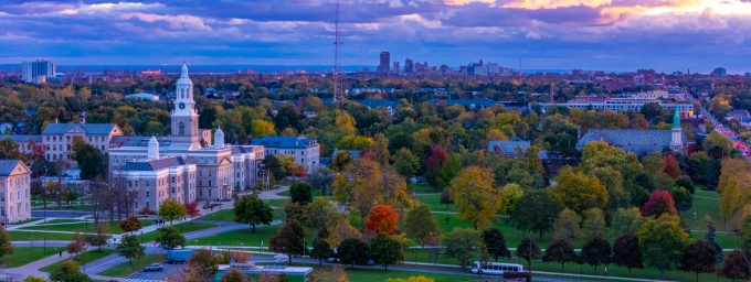 Aerial view of a campus and surrounding cityscape with historic buildings, autumn trees, and a colorful sunset sky.