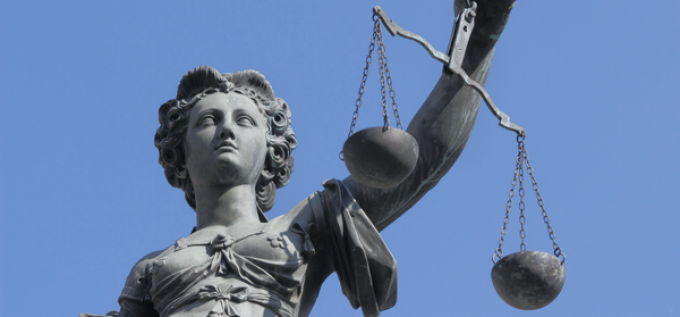 Statue of Lady Justice holding a set of scales in her right hand against a clear blue sky. The figure is depicted wearing a draped robe with detailed folds.