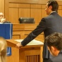 A courtroom where a young man at a podium addressing a judges bench where two men sit and listen. 