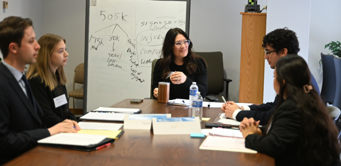 Five individuals seated around a rectangular conference table engaged in a mediation exercise. The table holds notebooks, folders, pens, and water bottles. A whiteboard in the background displays handwritten notes, including “505k” at the top with branching lines and words such as “injunction,” “comp,” and “limit.” An office cabinet and chairs are visible in the background.