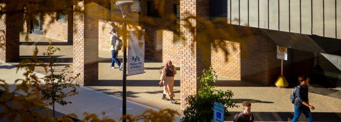 Students walking along a sidewalk next to a brick academic building on a sunny day. Yellow autumn leaves partially frame the view in the foreground. A banner with the University at Buffalo logo hangs from a lamppost near the building entrance.