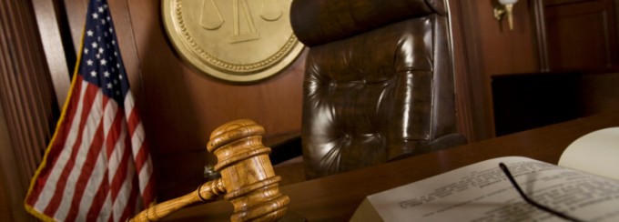 A low-angle view of a courtroom scene featuring a wooden judge’s bench with a leather chair behind it, a wooden gavel resting on the desk in the foreground, and an open book partially visible. An American flag stands to the side, and a circular wall emblem with scales of justice is mounted behind the chair.