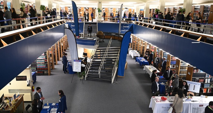 A large library, stairway in the center leding up to second floor, tables and lots of people standing around, talking. 