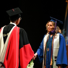 A graduate in a blue cap and gown shakes hands with an academic official in ceremonial robes on a stage during commencement. Flags stand in the background, and floral arrangements line the front of the stage.