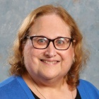 Woman wearing blue sweater, glasses, smiling. 