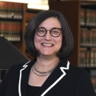 Woman wearing black suit with white trim, glasses, standing inside a library next to a table. 