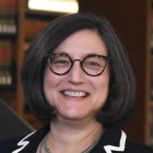 Woman wearing black suit with white trim, glasses, standing inside a library next to a table. 