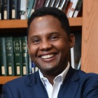 Man wearing suit, smiling, standing in front of a bookshelf in a library. 