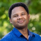 Man wearing blue shirt, smiling, standing outside near some trees. 