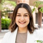 Woman smiling, standing outside in front of a brick building, next to some trees. 