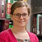 Woman wearing glasses, smiling, in a library next to a bookshelf. 