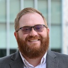 Man standing outside in front of a building, smiling, wearing glasses and suit. 