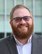 Man standing outside in front of a building, smiling, wearing glasses and suit. 