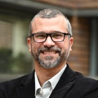 Man wearing glasses, smiling, standing outside in front of a brick building. 