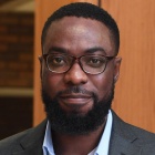 photo of man standing in front of a wooden all, wearing glasses and suit, smiling. 