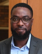 photo of man standing in front of a wooden all, wearing glasses and suit, smiling. 