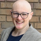 woman wearing glasses, smiling, standing outside next to a brick wall. 