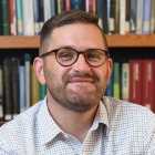 Man wearing glasses, smiling, sitting in front of a bookshelf with books. 