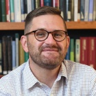 Man wearing glasses, smiling, sitting in front of a bookshelf with books.