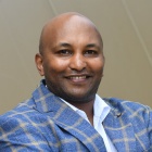 man smiling, wearing blue striped jacket, arms folded, standing outside. 